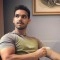 Man sitting in a room, looking to the side with a pensive expression. He wears a gray t-shirt and is seated casually. The background features a framed artwork on a beige wall.