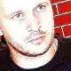 Close-up of a man with a beard in front of a brick wall, looking to the side. The image has high contrast, emphasizing facial features. He wears a dark t-shirt.